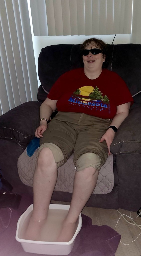 Nickiesoaks her feet in a tub of warm water. Her khakis are rolled up to her knees. She wears a red Minnesota print t-shirt.