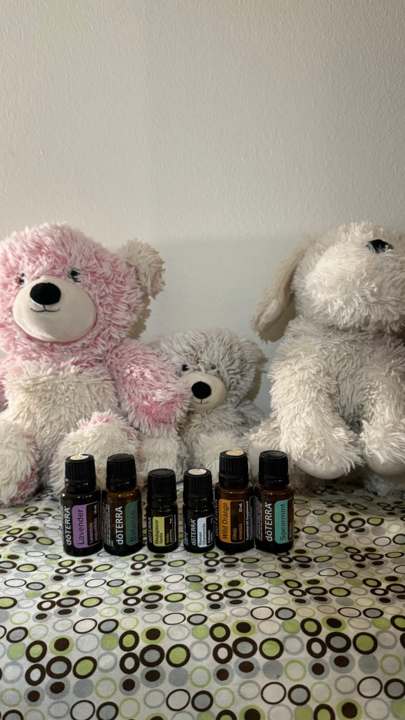 Three Warmie stuffed animals appear to be holding bottles of essential oils. From left to right, they are a pink Marshmallow bear Warmie holding Lavender and Balance, a white Marshmallow bear Warmies Junior holding Madagascar Vanilla and Roman Chamomile and a yellow lab Warmie holding Wild Orange and Spearmint 