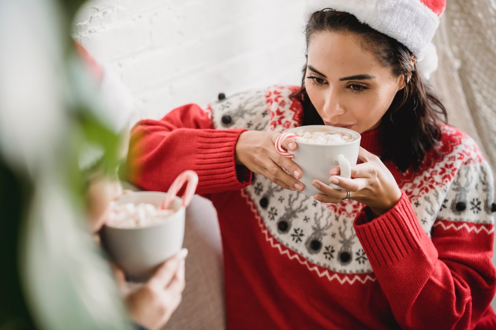 A woman wearing a red and white Christmas sweater holds a mug of hot chocolate with marshmallows and a candy cane.  