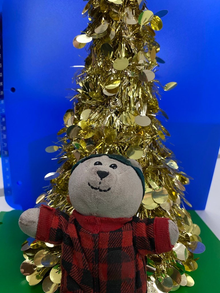 A 4 inch teddy bear wearing red Christmas pjs and sleep shade sits propped against a tabletop Christmas tree in front of a blue background. 