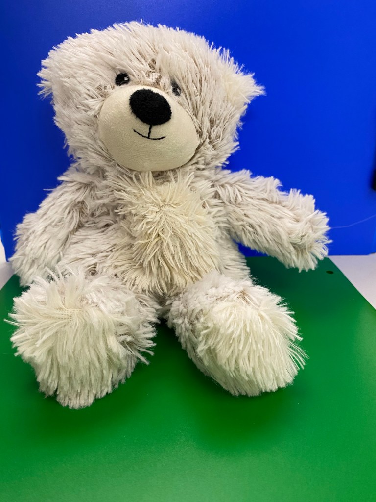 A fluffy white teddy bear sitting on a green surface with a blue background. The teddy bear has a friendly face with a black nose and eyes, and its fur appears soft and shaggy. It has its arms and legs spread out, giving it a relaxed and inviting posture.