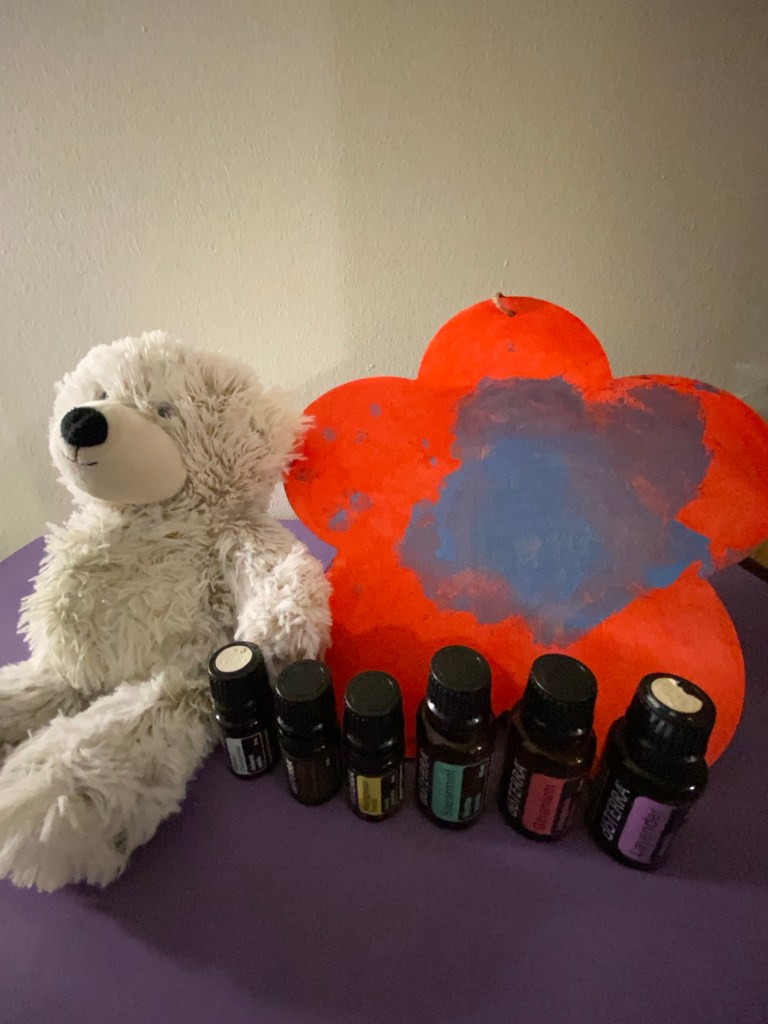 A white teddy bear sits beside a red flower-shaped painting with a blue center on a purple surface. In front of them, 6 doTERRA essential oil bottles are lined up. The essential oils are Geranium, Roman Chamomile, Lavender, Sandalwood, Madagascar Vanilla and Spearmint.